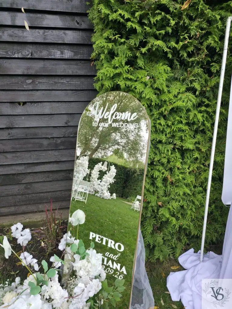 Дзеркало «Welcome» біля входу Арочне дзеркало «Welcome to our wedding» з білим написом біля зони церемонії, внизу композиція з білих квітів